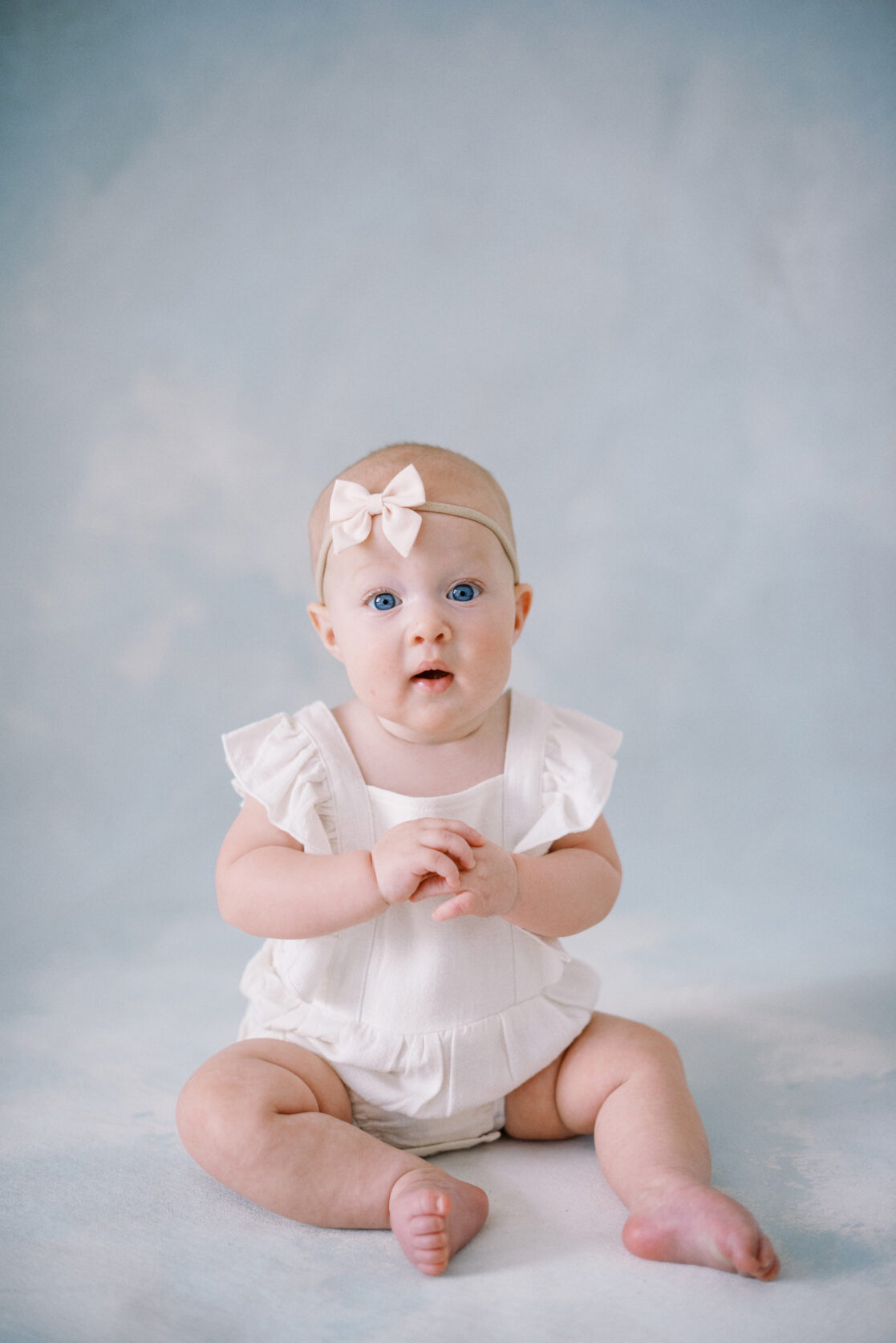 Photo of a baby girl wearing cute baby clothes in Richmond against a watercolor blue backdrop wearing a white frilly romper with a light pink  bow on her head. 