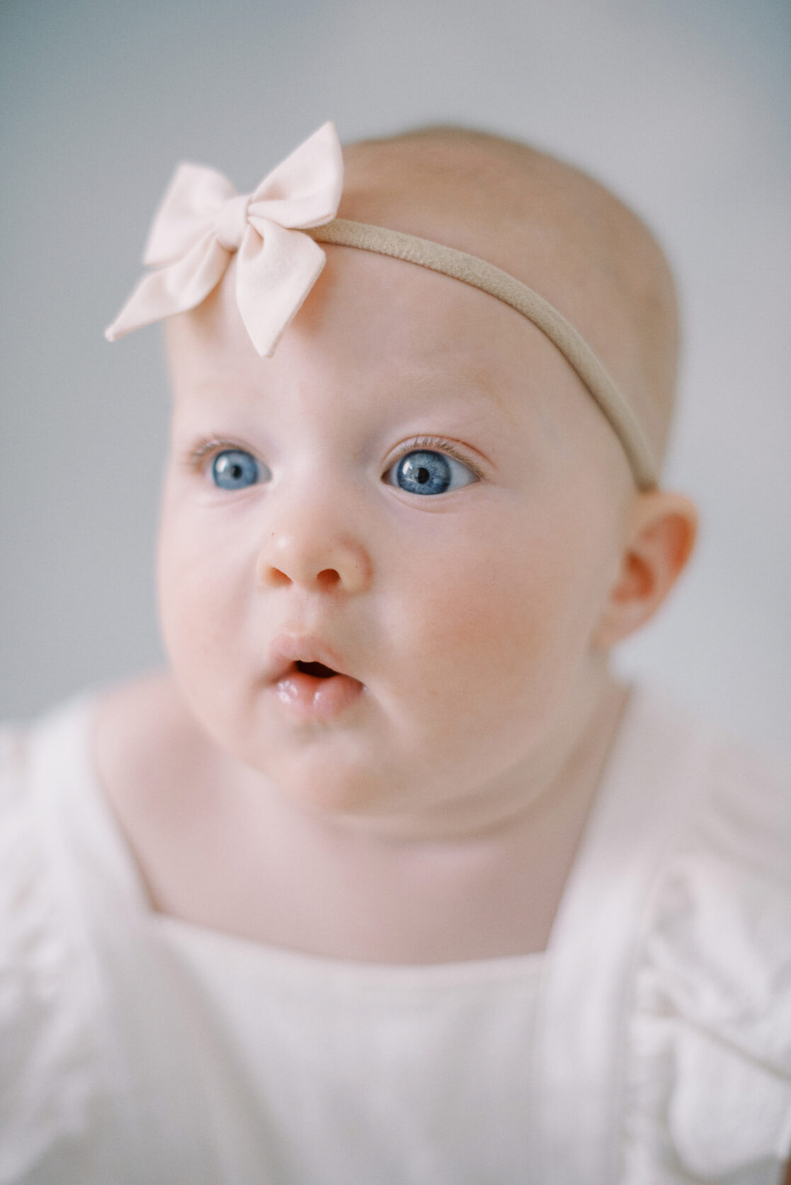 Photo of a baby girl wearing cute baby clothes in Richmond against a watercolor blue backdrop wearing a white frilly romper with a light pink  bow on her head. 