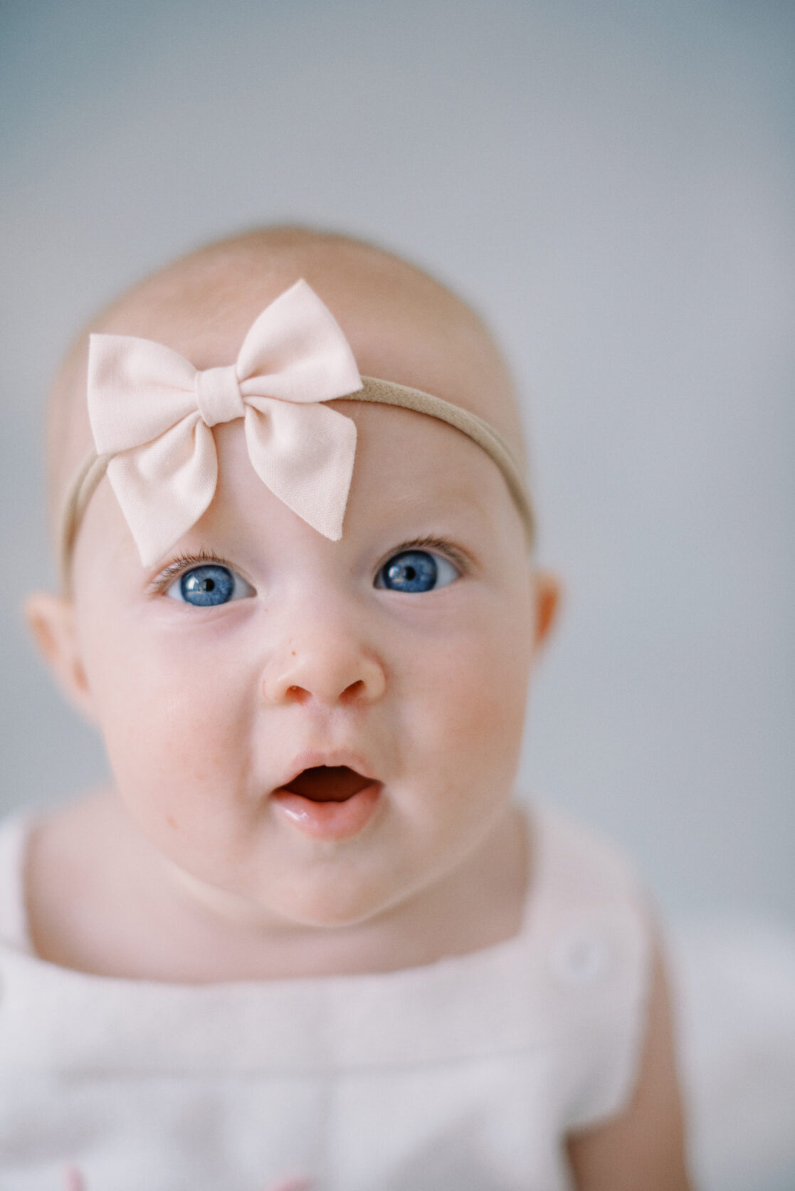 Photo of a baby girl wearing cute baby clothes in Richmond against a watercolor blue backdrop wearing a white frilly romper with a light pink  bow on her head. 