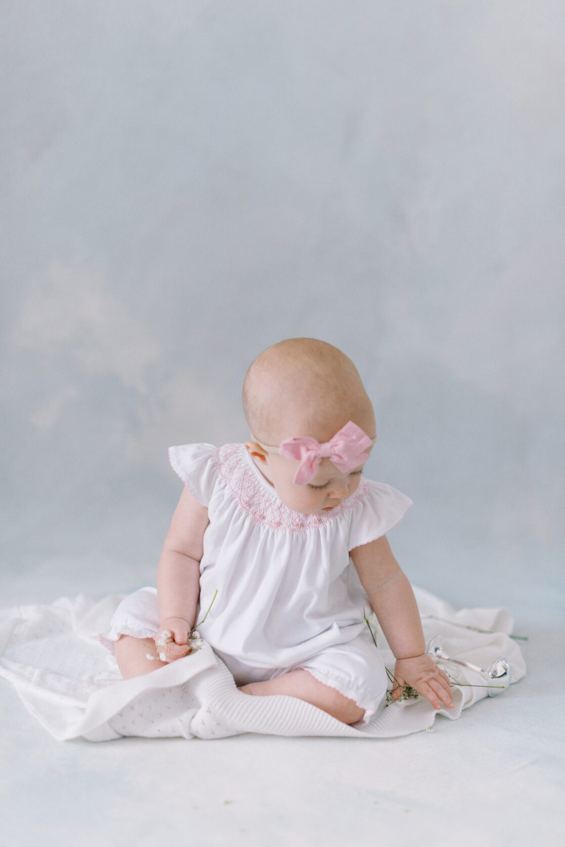 Photo of a baby girl wearing cute baby clothes in Richmond against a watercolor blue backdrop wearing a white frilly romper with a light pink  bow on her head. 