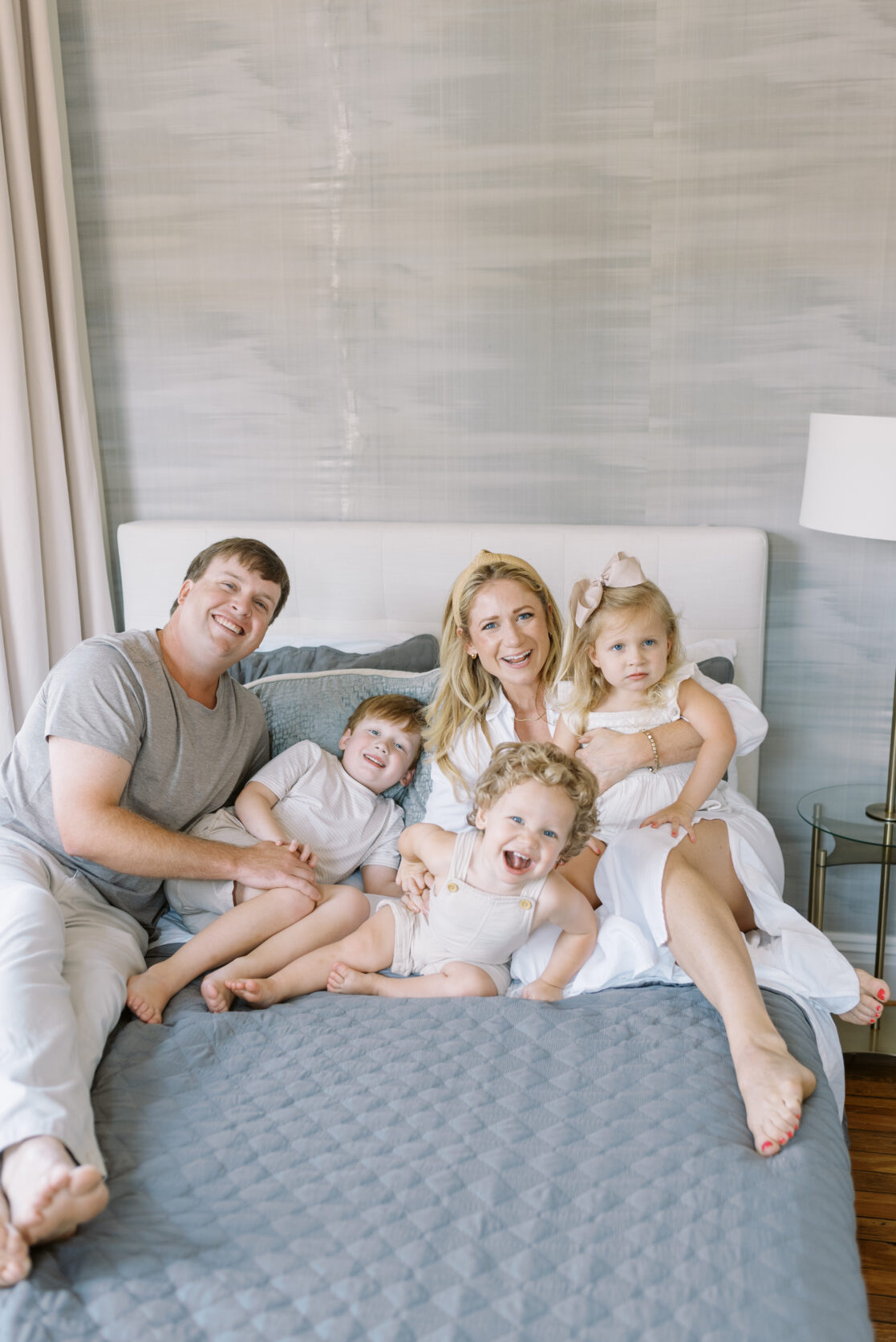 Mom and dad with three children in coordinating beige and white outfits laying on a bed in brightly lit bedroom during fun family photos in Richmond by Jacqueline Aimee Portraits 