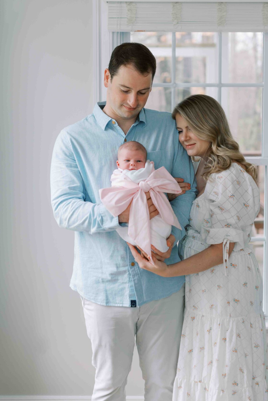 Photo of a two parents, a mom and a dad, holding their newborn daughter wrapped in a white blanket and pink bow. The parents booked their newborn photos at 10 days old, a great time if you're wondering when to schedule newborn photos 