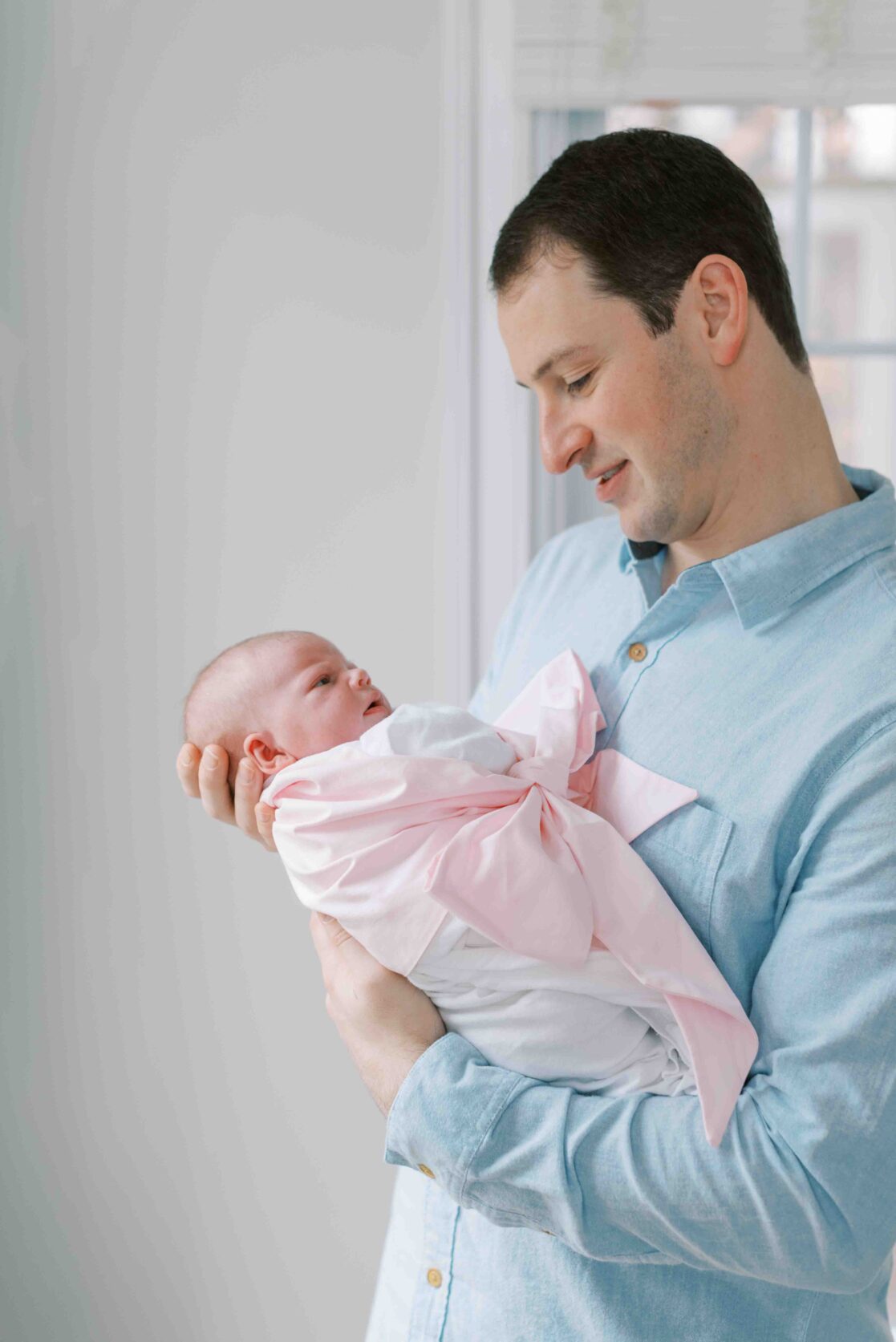 A dad in a blue shirt holds his newborn daughter wrapped in a white blanket and pink bow. The parents booked their newborn photos at 10 days old, a great time if you're wondering when to schedule newborn photos 