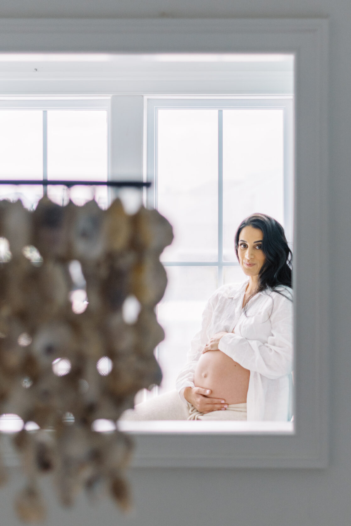 Richmond Birth Doula | Photo of a pregnant mom holding her belly in the background and looking at the camera. In the foreground is an out of focus oyster chandelier by Richmond maternity photographer Jacqueline Aimee Portraits