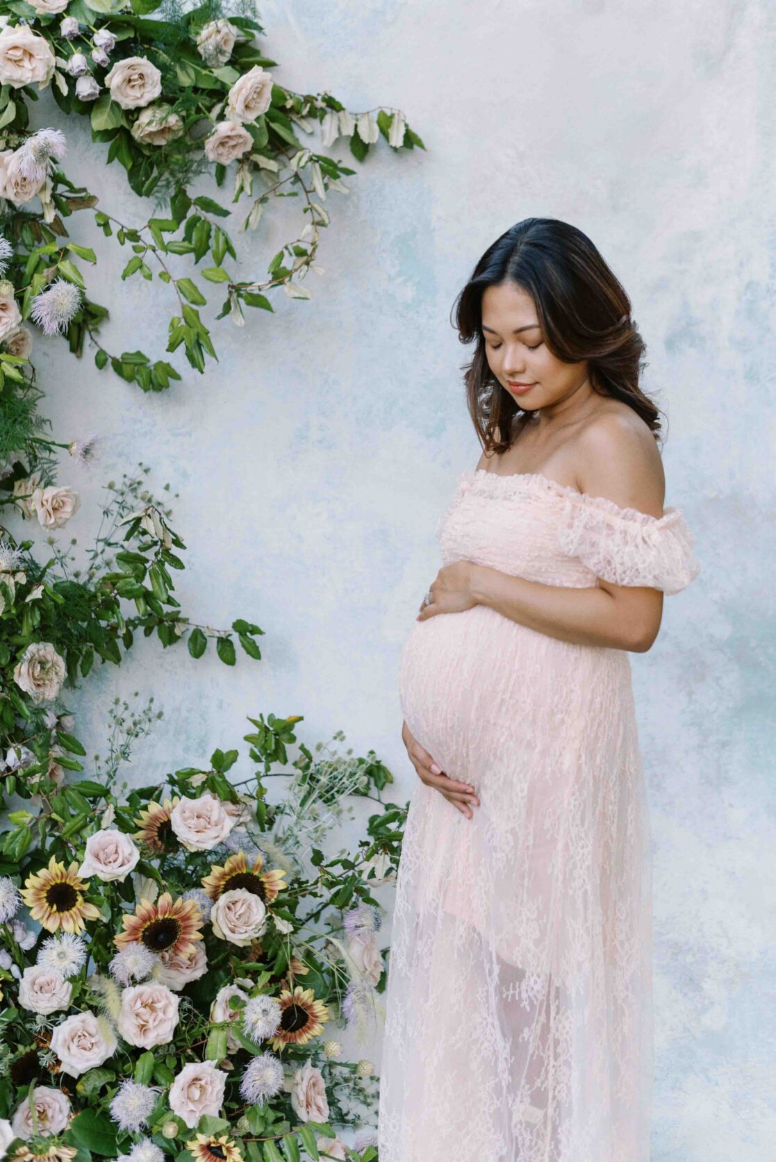Richmond birth doula | Photo of a pregnant mom holding her belly in a pink lace dress, looking down towards her growing belly. She is in front of a lovely watercolor backdrop and floral arrangement by Richmond maternity photographer Jacqueline Aimee Portraits